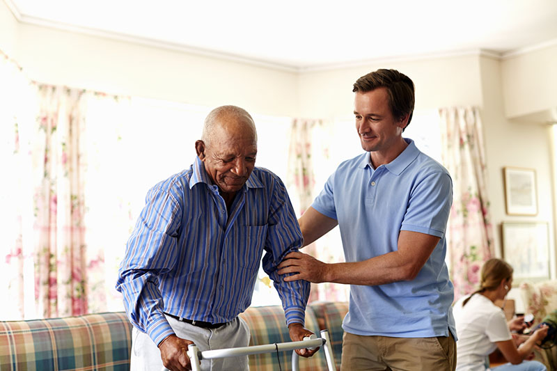 Male caretaker assisting senior man with walker in nursing home